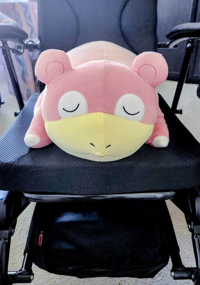 A front view of a powerchair with a black frame and black cushions, with a "slowpoke" sleeping pokemon plush sitting in the chair.