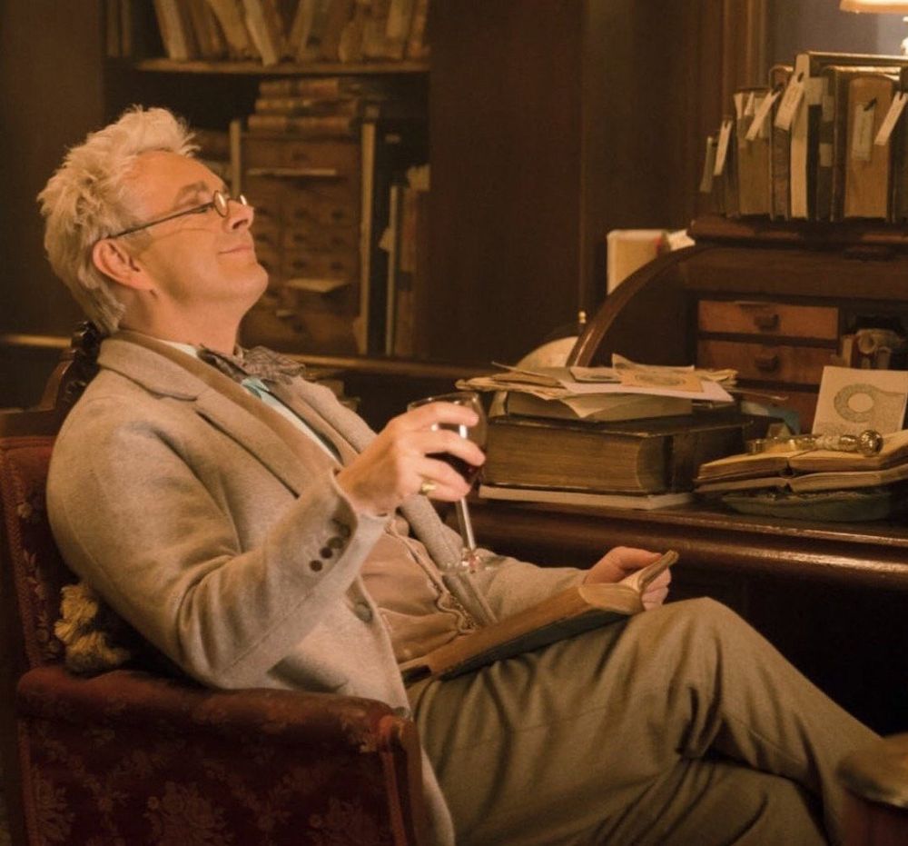 Michael Sheen as Aziraphale in Good Omens. His hair is light blonde and fluffy, and he is sitting in the bookshop, reclining in a chair with a glass of wine and a book, his glasses, and his eyes closed. 