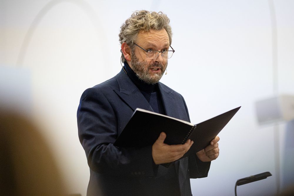 Michael Sheen at the Burton100 Gala. He is wearing a dark blazer jacket with a turtleneck (!!!) and his glasses, and performing while holding a black notebook