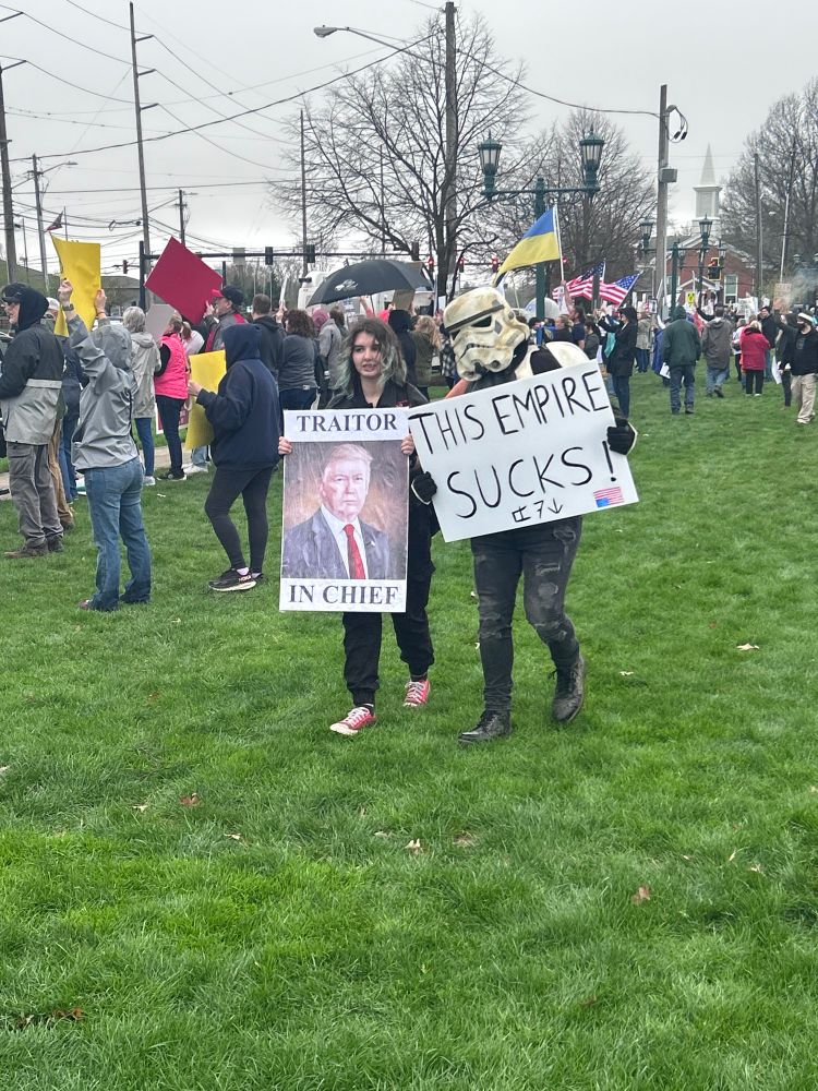 Two protestors one carrying a picture of Trump captioned “Traitor in Chief.” The other in a Stormtrooper from Star Wars mask and a sign reading “This empire sucks.”