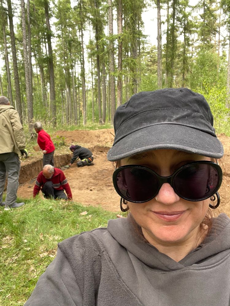 A selfie of a white woman wearing a black cap, huge black sunglasses and a grey hoodie, standing in a forest, with an ongoing excavation trench in the background. 