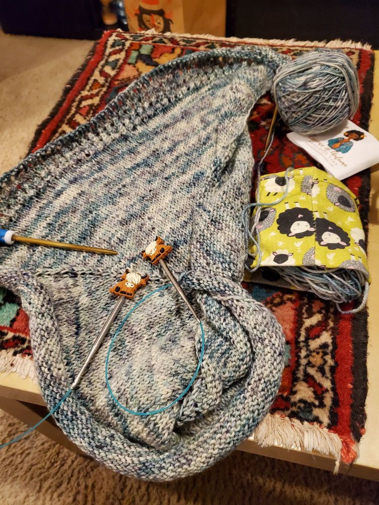 A green knitted shawl in progress rests on a Persian-style rug atop a coffee table. 