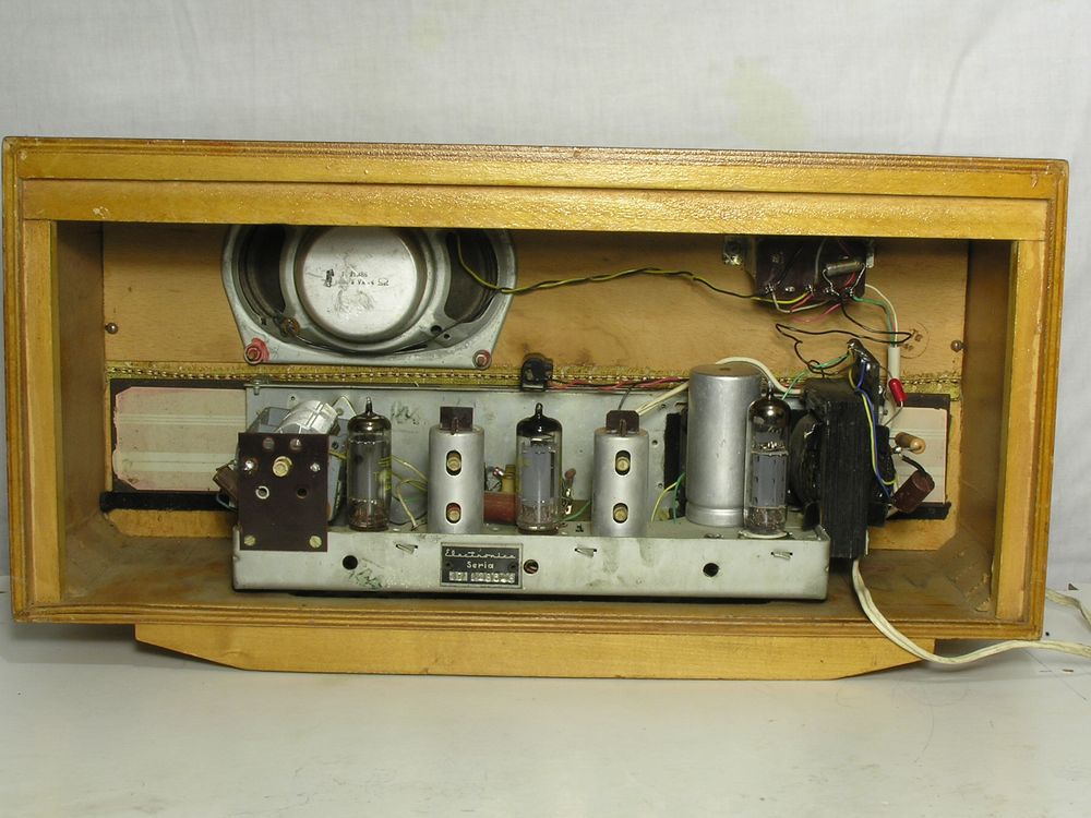 The same radio as before, now with the back cover removed.  A small elliptical speaker is visible along with the audio output transformer, both screwed to the speaker panel.  Below these, a metal chassis on which is the variable capacitor for tuning, three valves, two cylindrical IF transformers, a cylindrical electrolytic capacitor (for smoothing the HT supply).  Hanging off the right of the chassis is the mains transformer providing the HT and heater supplies, but not providing mains isolation.