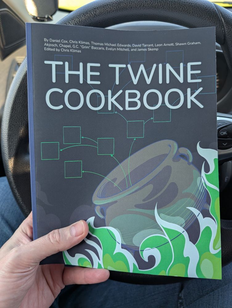 Picture showing a physical book, The Twine Cookbook, held in the hand of a person sitting in the driver's seat of a car with the steering wheel behind the book.