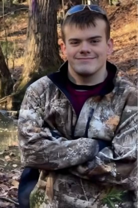 Phoenix Ikner (FKA Christian Gunnar Eriksen) poses for a photo. He is wearing a camouflage patterned jacket, has his arms crossed in front, and is smiling with a closed mouth. 