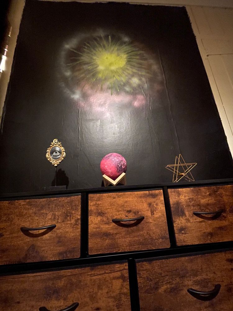 A small dark chesf of drawer with a planet, a star and an eye as ornaments on top. The wall behinds is black but has some abstract bright paint suggesting stars or space. 