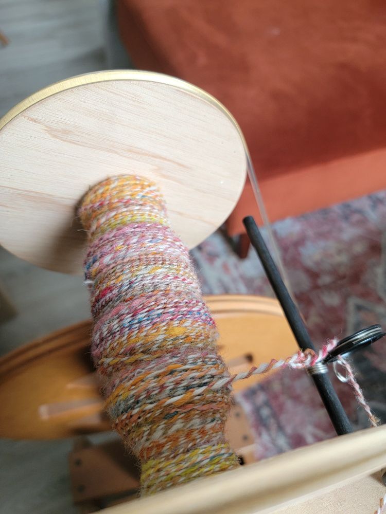 A spinning wheel from above, showcasing yarn being spun in shades of yellow, pink, orange, blue, and red. The colourful yarn is plied with a strand of white yarn. 