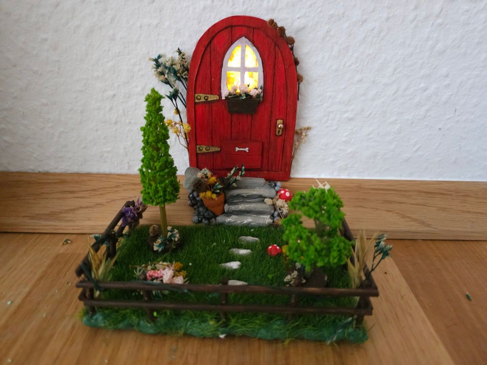 Foto einer kleinen roten Wichteltür mit aufwändigem Garten. Aus dem keinen Fenster kommt Licht. 