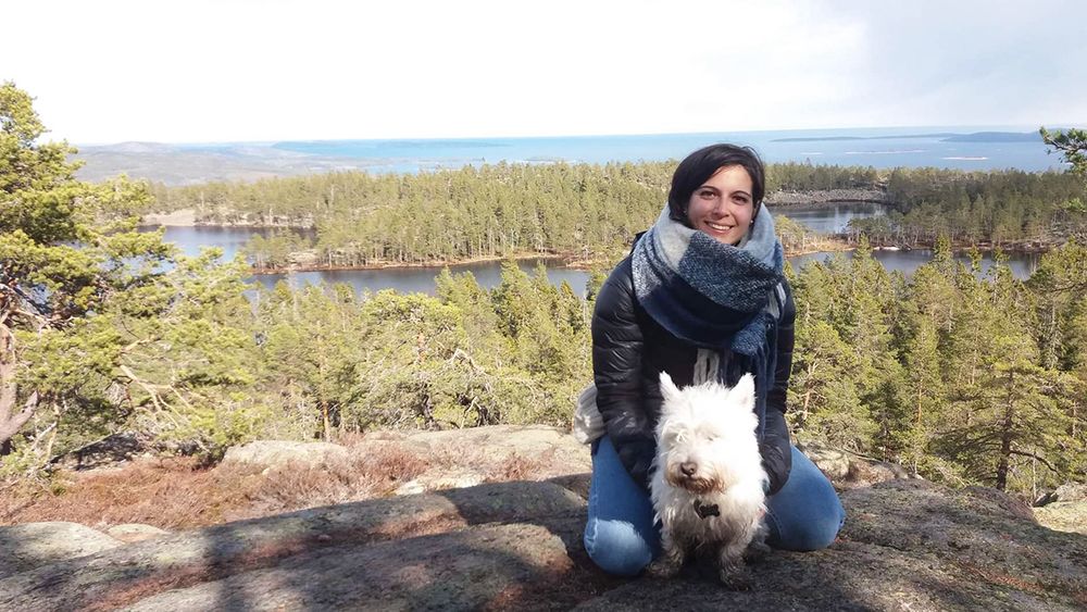 Alice Marcon is sitting on a stony hill in front of a forest landscape that goes over to the sea. In front of her is sitting a small white dog. She has black hair, a scarf around her neck, jeans and a jacket on and is smiling into the camera. 