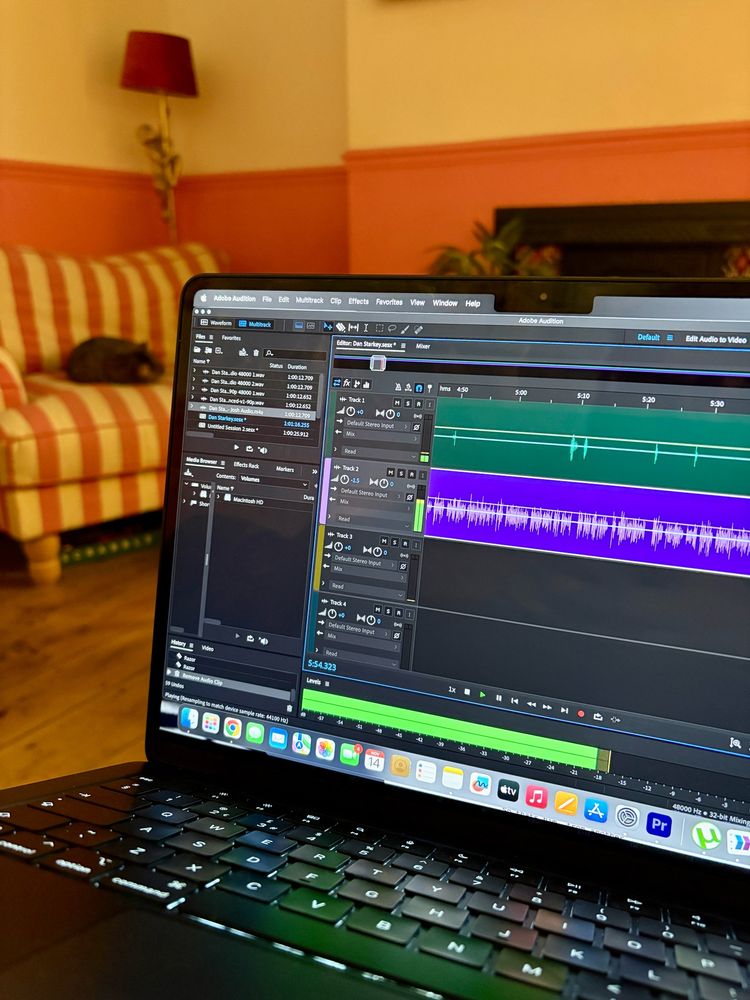 A photo of a laptop showing an in progress podcast edit on Adobe Audition for an upcoming episode of Who Knew?, with my cat on a chair in the background!