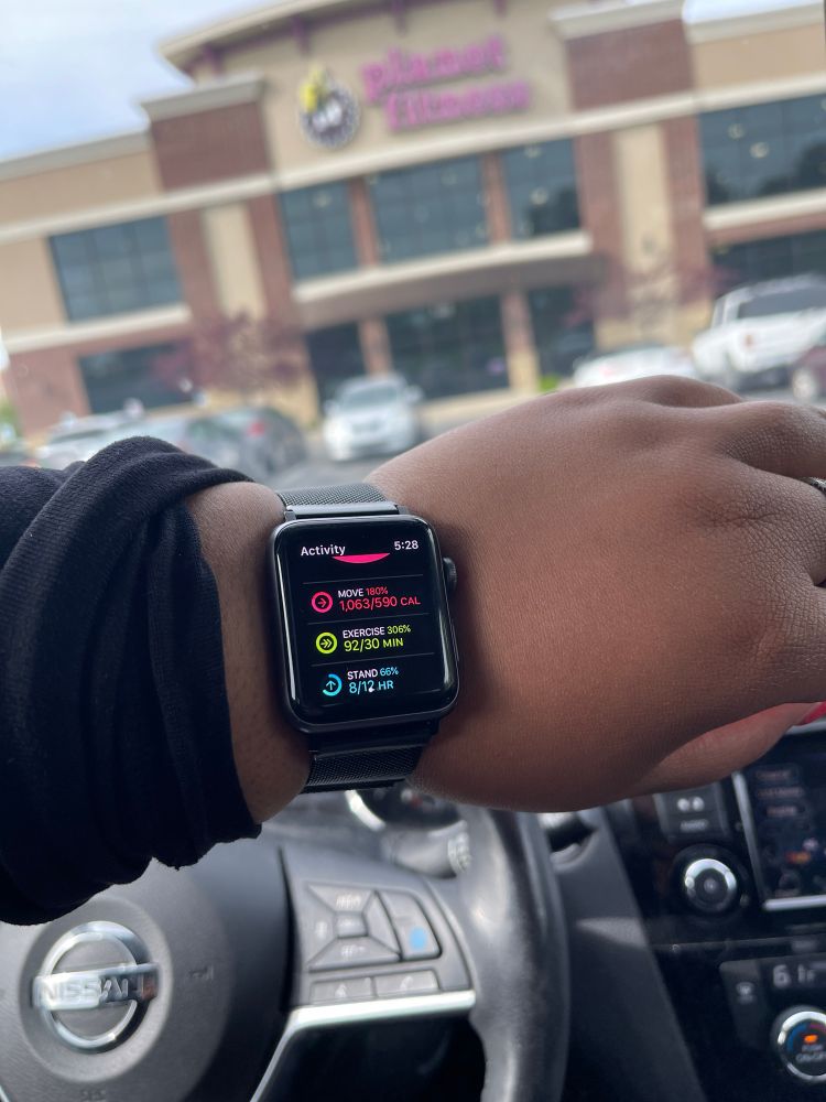 Tanesha’s rest displaying her Apple Watch. It shows that she has burned 1063 cal, has exercise for 92 minutes, stood for 8 hours so far. In the background is Planet Fitness and the parking lot.