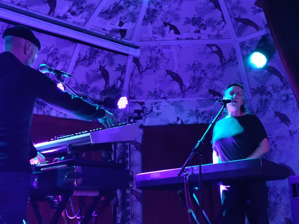 Picture taken from front left of stage. Lomond Campbell left, Kathryn Joseph right, facing each other over their keyboards (his synths, hers piano) with his back towards us. They are lit by  blue spotlights against a purple background of the legendary Deaf Institute wallpaper.