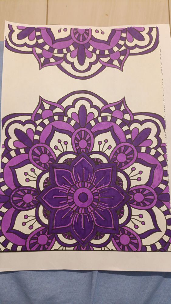 Na imagem apresenta uma folha branca com uma andala em formato florido, a mandala foi pintada em um tom de roxo claro e outro tom de roxo mais escuro.