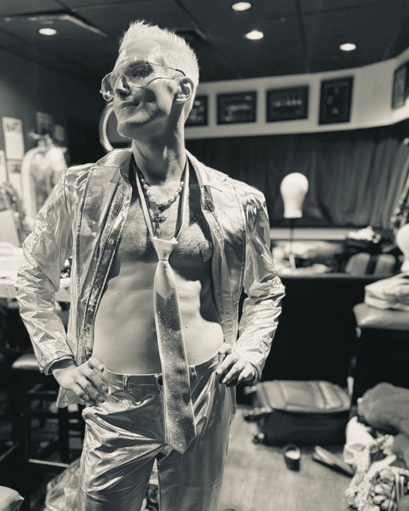 A black and white photo of a blonde guy with hands on his hips, open shirt, a bright tie, wearing glasses, a bright silver pants 