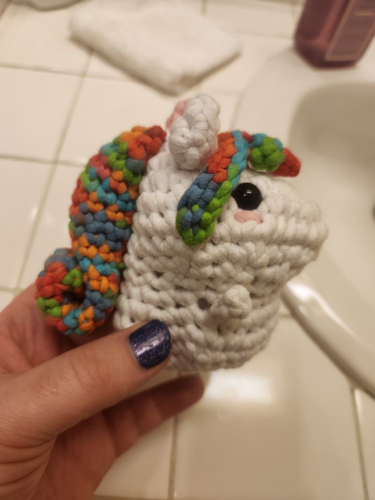 A white yarn crochet unicorn with a rainbow main and bangs. It has arms, ears, and a pink horn though it's not easy to see in this shot. Its blushing pink