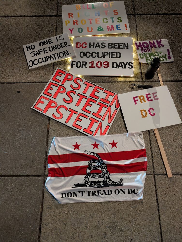Protest materials laid out on a sidewalk at night. Visible signs read: "BILL OF RIGHTS PROTECTS YOU & ME!," "NO ONE IS SAFE UNDER OCCUPATION," "DC HAS BEEN OCCUPIED FOR 109 DAYS," "HONK FOR DEMOCRACY!," "EPSTEIN EPSTEIN EPSTEIN," and "FREE DC." A black water bottle and a spoon sit near the signs. At the bottom is a DC flag-style banner with red bars, three stars, a coiled snake, and the text "DON’T TREAD ON DC."
