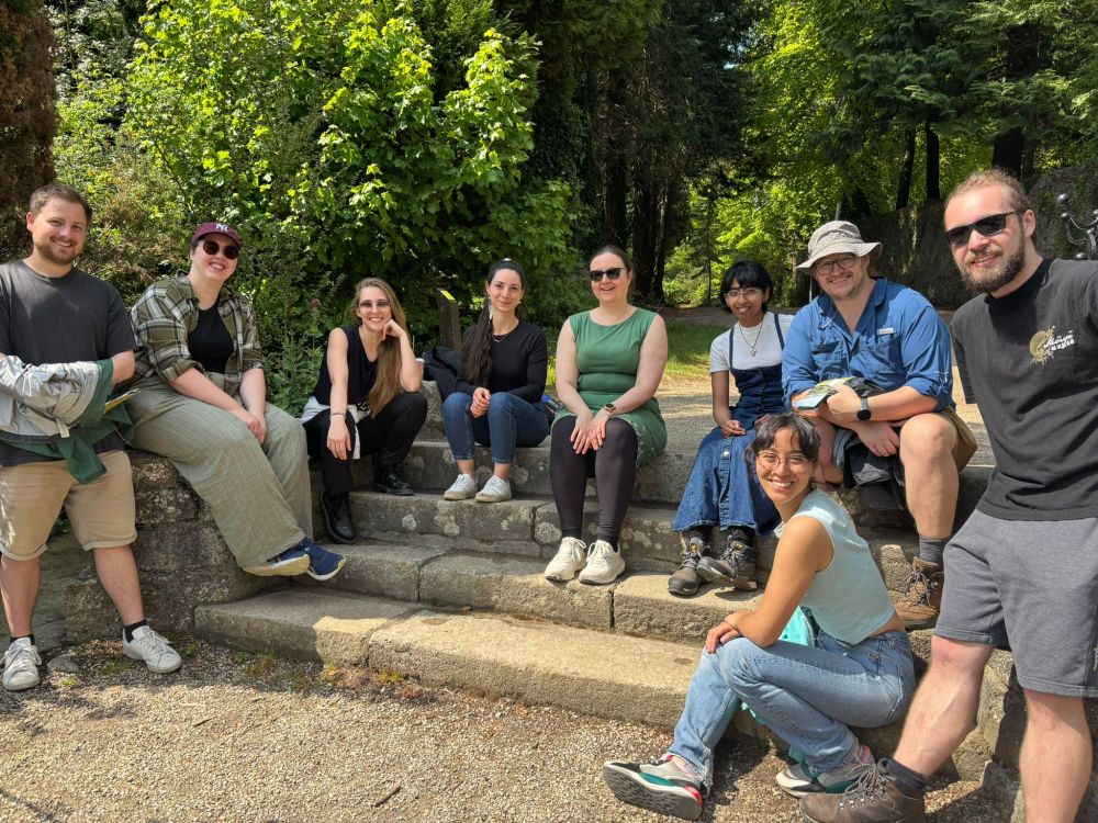 Nine people in summer clothes and sunglasses, smiling, sitting on stone steps, with trees in the background