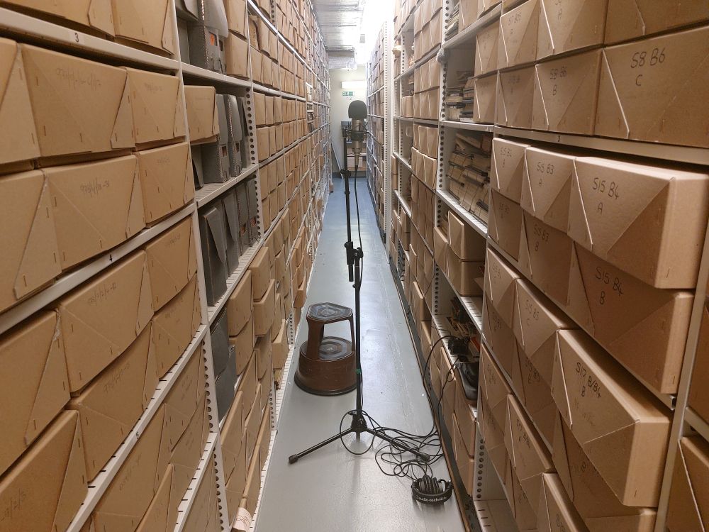 A microphone stand set up in the far aisle of an archive strongroom. Acid-free archive boxes are on the shelves. A kickstool is in the aisle behind the stand.