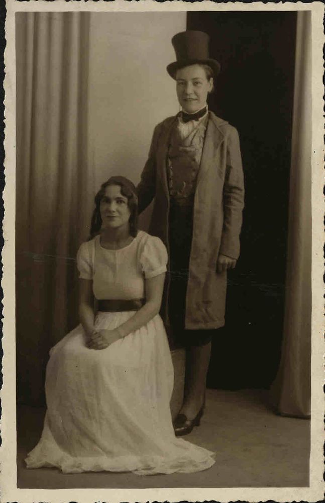 Late 1920s or early 1920s photograph of two women. One (Eileen Younghusband) is wearing a white dress and sitting down. The other (identity currently unknown) is standing behind her and resting one hand on the back of the chair, she is wearing a top hat, bow tie, waistcoat and frock coat.