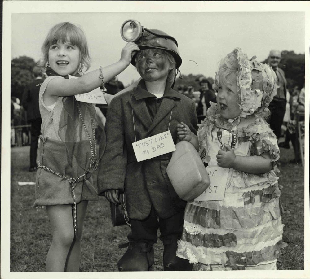 Photograph of children at 1969 Yorkshire Miners' Annual Demonstration, held in Barnsley. The three children have entered the fancy dress competition. The boy in the middle is dressed as a miner, complete with hat, bottle and sign saying 'Just like mi dad'.