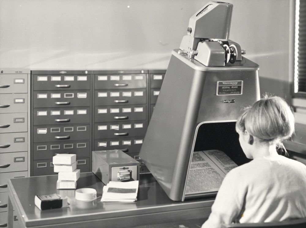 Black and white photograph taken in the University of Warwick library in the 1960s. It shows a woman looking at microfilm on the 'Archival Reader', a big manual microfilm reader. Metal filing cabinets containing rolls of film can be seen in the background.