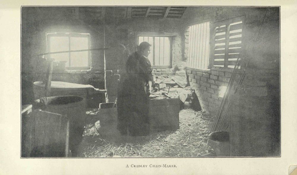 Printed black and white photograph of a young woman with her back to the camera, working at a forge with a small child facing the camera looking on.