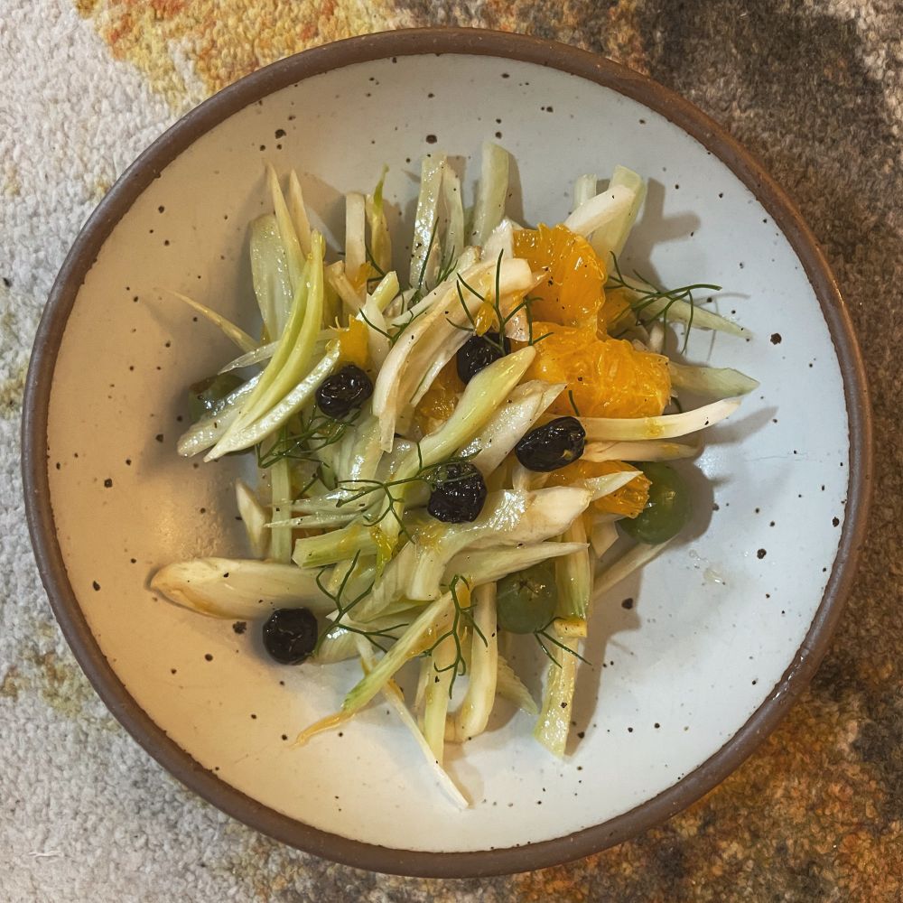 An eggshell-colored bowl from East Fork filled with slices of fennel, onion, navel orange, plus castelvetrano olives, dried sour cherries, and fennel fronds. 