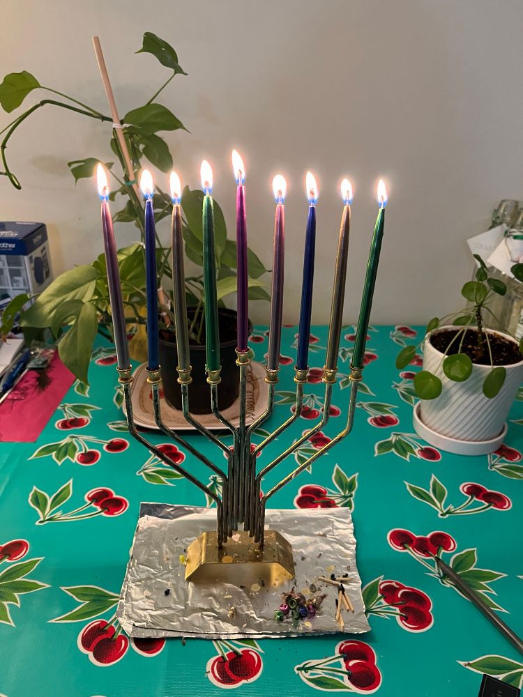 9 candles burn in a tall silver and gold menorah on a table covered in red cherry print oilcloth with plants in the background. The monies rests on ritual tinfoil, with spent Gramercy Tavern matches, as G-d intended. 