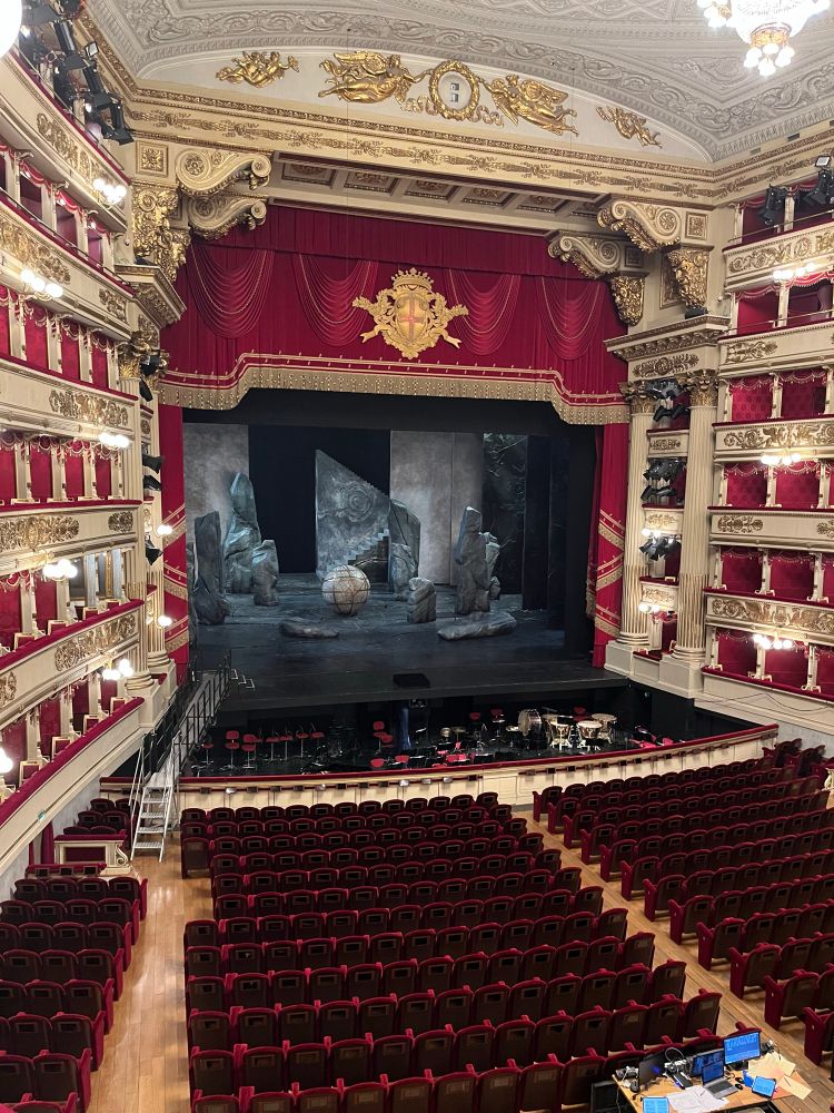 La Scala interior during a break from a rehearsal of Die Walkurie. They are adjusting the lighting and the stage has big grey rocks and a sphere. The theater is lit and you can see the red velvet orchestra seating. 