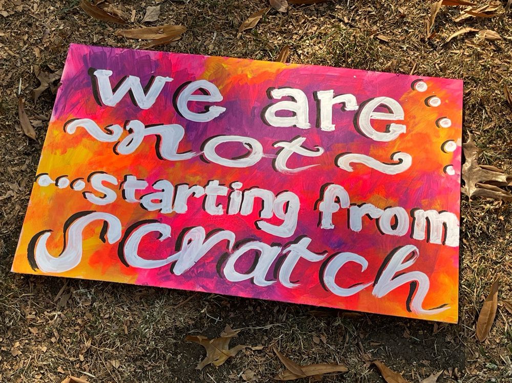 Yellow, orange, and purple tie-dye-ish sign that says “we are not starting from scratch” lying on the grass