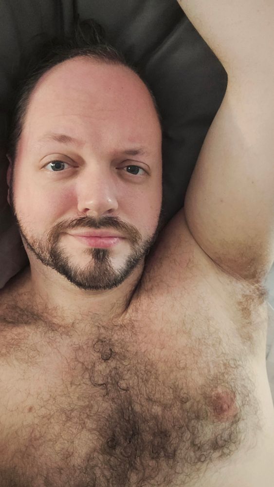 Wessl shirtless in bed with an arm raised over their head