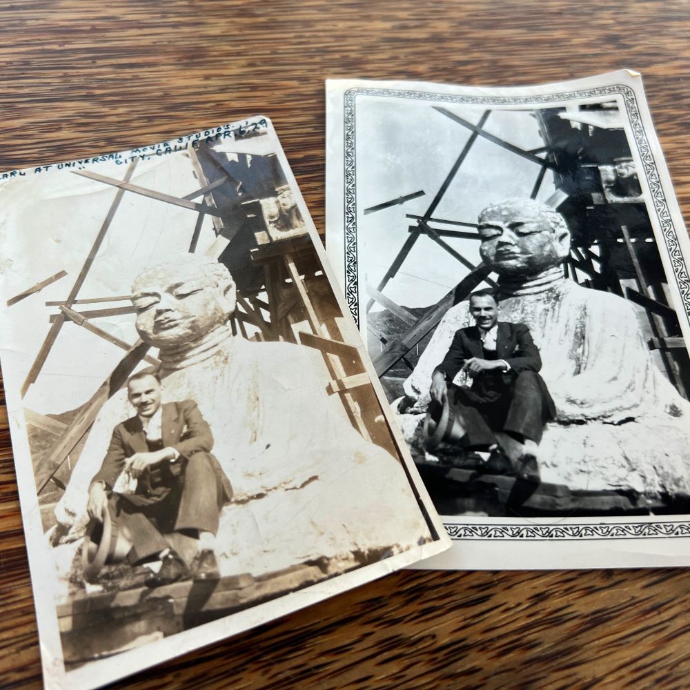 The image features two black-and-white photographs placed on a wooden surface. Both photographs depict a man in a suit sitting beside a large sculpture of a seated figure, possibly a Buddha, surrounded by a scaffolding structure. The photograph on the left is slightly sepia-toned and shows the man sitting on the base of the statue, holding an object. The photograph on the right is clearer with a decorative border, showing a similar scene with the man in a slightly different pose. The statue appears unfinished or under construction with visible scaffolding.