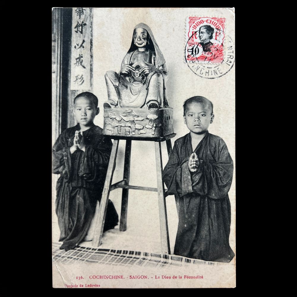 The image is a vintage postcard featuring a statue of a seated icon, placed on a wooden stand. The statue depicts a smiling figure in a robe, with its hands holding open his chest to reveal a face. Flanking the statue are two young novice monks in dark robes, standing on either side with hands held in prayer. A visible red postage stamp and markings are at the top right corner of the image, the cancellation date of 1911 is barely visible. 
Sort available posts on Bluesky using #Buddhasinthewest | All posts archived here: tinyurl.com/4kmcwp87 🔍 🗃️ 📜
