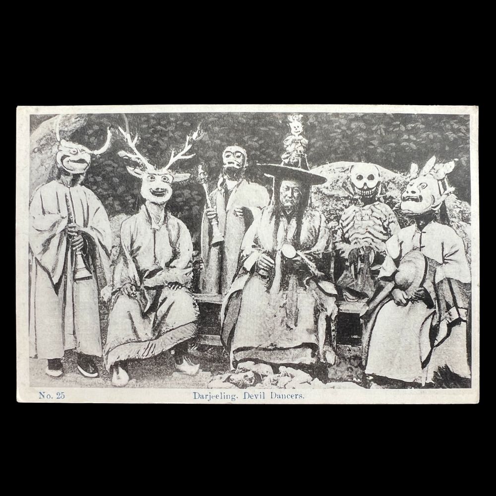 The image is a black and white photographic postcard of six individuals dressed in elaborate, ceremonial costumes. Each person is wearing a distinct mask with animal and demonic features. On the far left, a figure wears a cloak and a bird mask with horns, holding a Tibetan-style trumpet. Next, a seated person dons a mask resembling a deer with large antlers. Beside them stands another individual holding a Tibetan-style trumpet. The central figure is seated, wearing a large, black hat with figuring attached to the brim. To their right stands a person with a skeleton-like costume, featuring a skull mask and rib-like decorations. The last figure on the right has a Mahakala demon-like mask, holding cymbals. The figures are posed in a natural outdoor setting.

Sort available posts on Bluesky using #Buddhasinthewest | All posts archived here: tinyurl.com/4kmcwp87 🔍 🗃️ 📜
