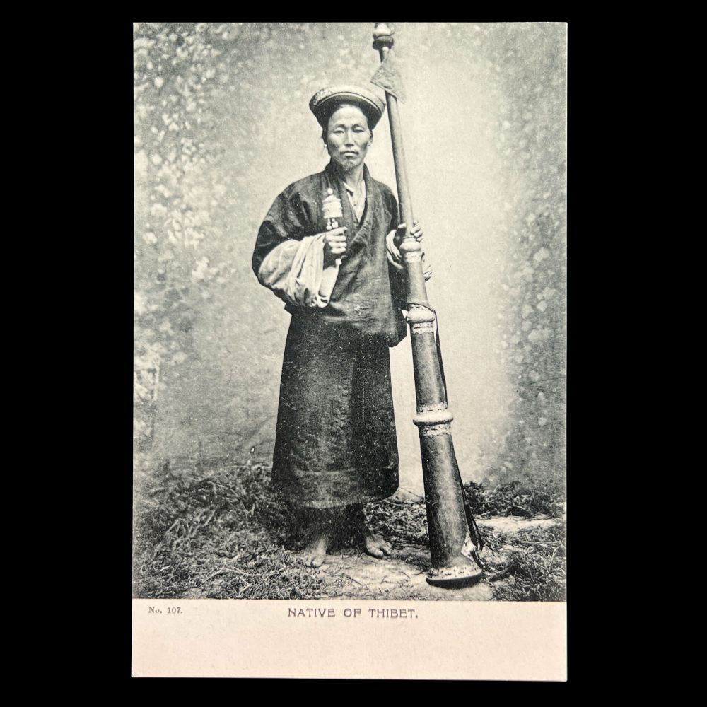 The image shows a black and white photograpic postcard of a person standing and holding a long conical horn taller than their height, and a hand-held Buddhist prayer wheel. The individual is dressed in a long robe and a head covering. The person stands on grass with a neutral, slightly dappled backdrop that suggests a studio setting. The individual is barefoot, and their expression is calm. The caption says, "Native of Thibet."