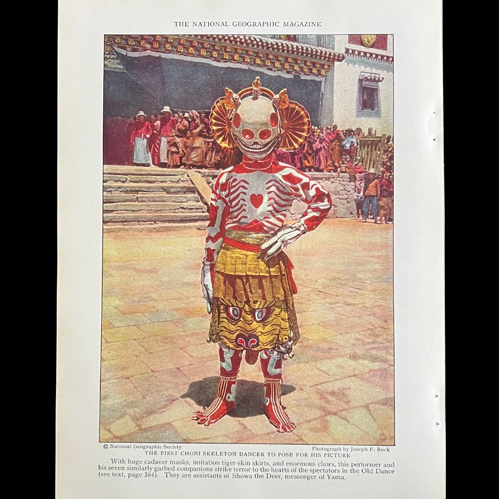 The image shows a performer in a traditional costume posing outdoors. The person is dressed in a vibrant and intricate costume resembling a skeleton, featuring red and white patterns over the body. The headpiece is a large mask with skeletal features, including a prominent circular face and exaggerated eyes. The costume includes a yellow and red skirt intended to depict a tiger skin. The individual stands on a stone-paved surface with one hand on their hip. Behind them, a group of monastics sit in the courtyard.