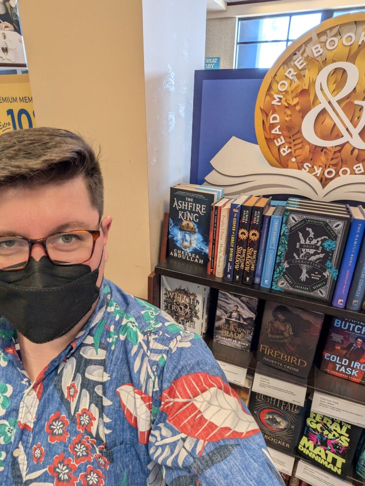 A selfie of me (Alex) standing next to a Barnes & Noble display shelf.
The visible book covers are The Ashfire King by Chelsea Abdullah, A Drop of Corruption by Robert Jackson Bennett, The Witch who Trades with Death, The Fireborne Blade by Charlotte Bond, Firebird by Juliette Cross, Cold Iron Task by James Butcher, Lightfall by Ed Crocker, and The Gate of the Feral Gods by Matt Dinniman.
I'm wearing a blue Aloha shirt patterned with various red flowers and green leaves, as well as a black KN95 mask below my tortoiseshell glasses as I smirk slightly with my eyes. 