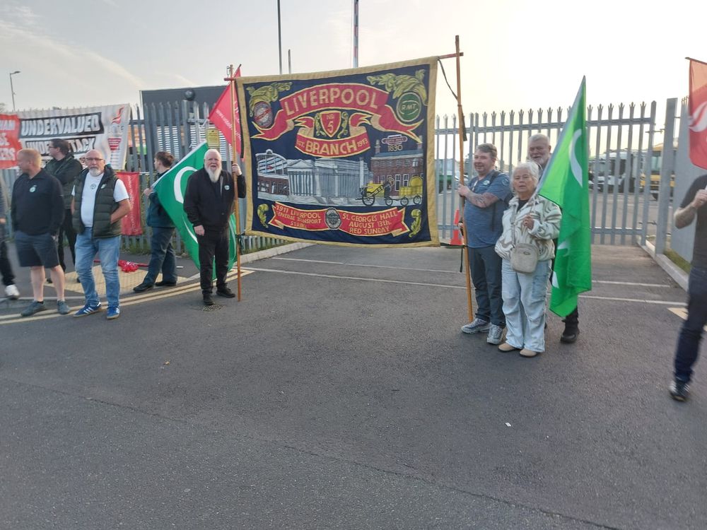 Group of people standing outdoors holding a large Liverpool Branch banner with detailed embroidery and green flags on a paved area near a metal fence.