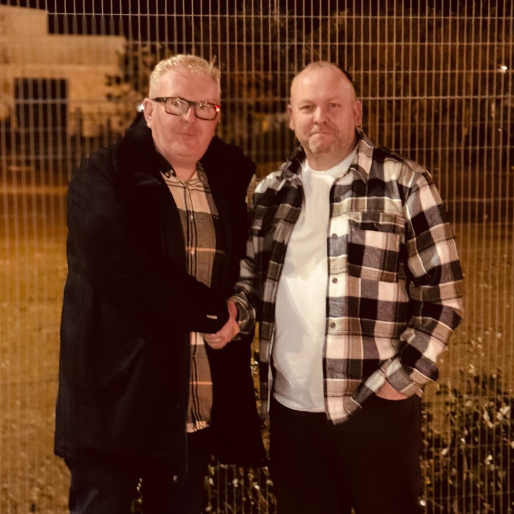 Two men shaking hands outdoors at night in front of a metal fence, both wearing casual jackets and plaid shirts.