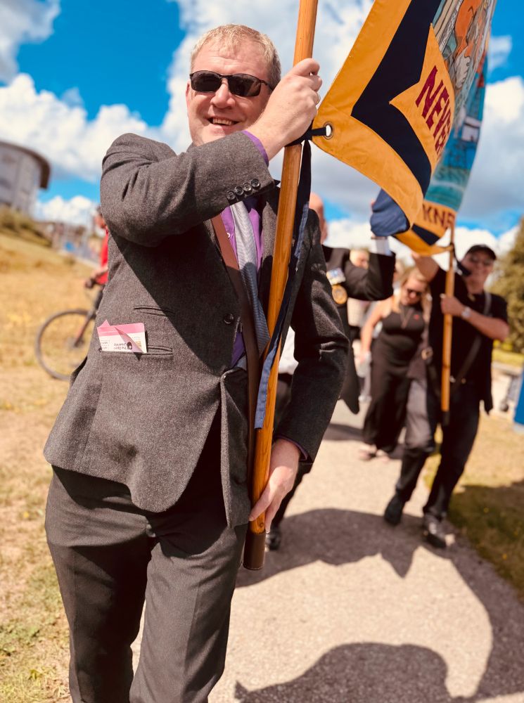 Person in a grey suit holding a wooden pole with a colourful flag during an outdoor event on a sunny day.