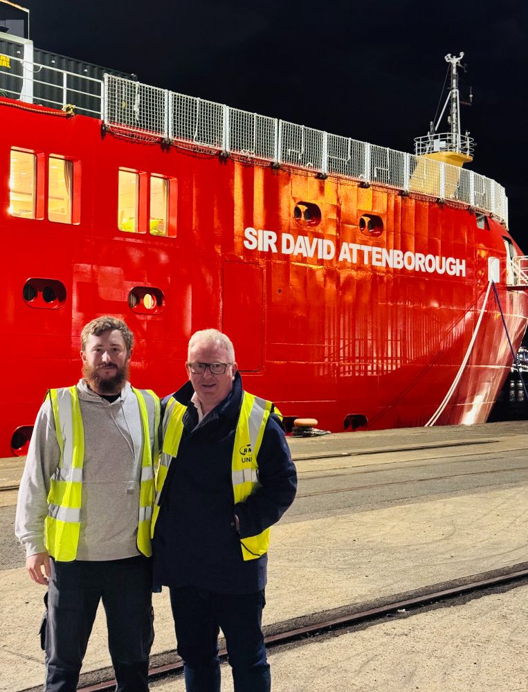 Two men wearing high-visibility vests stand on a dock beside the large red ship named Sir David Attenborough at night.