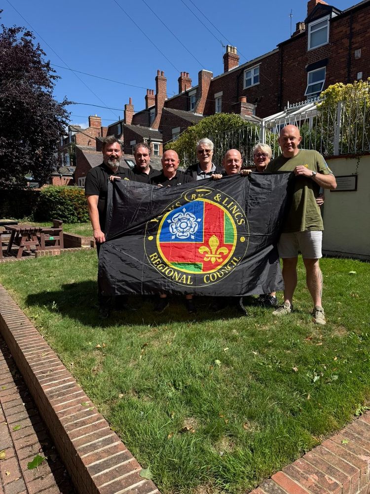 Group of seven people standing on a grassy lawn holding a black flag with a colourful emblem and text reading 'Yorks & Lincs Regional Council'.