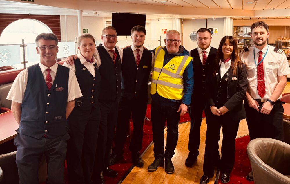 Group of eight crew members in uniform posing inside a ship's lounge with wooden floors and large windows.