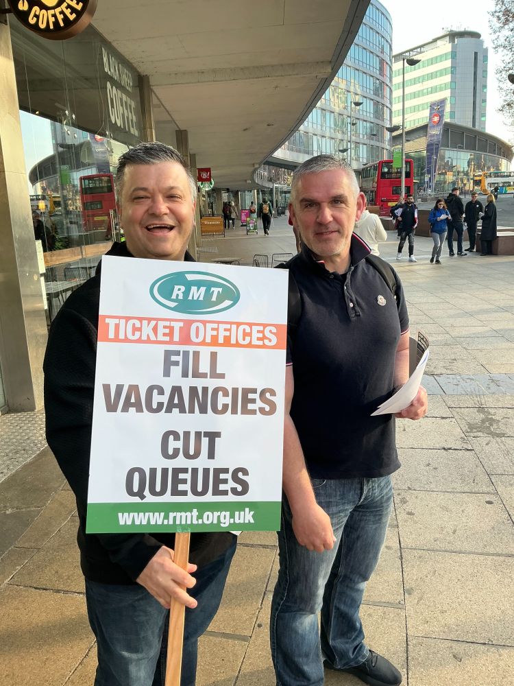 Two RMT members outside Manchester Piccadilly station smile for the camera. One holds leaflets whilst the other holds up a sign that reads 'Ticket Offices: Fill Vacancies, Cut Queues'.