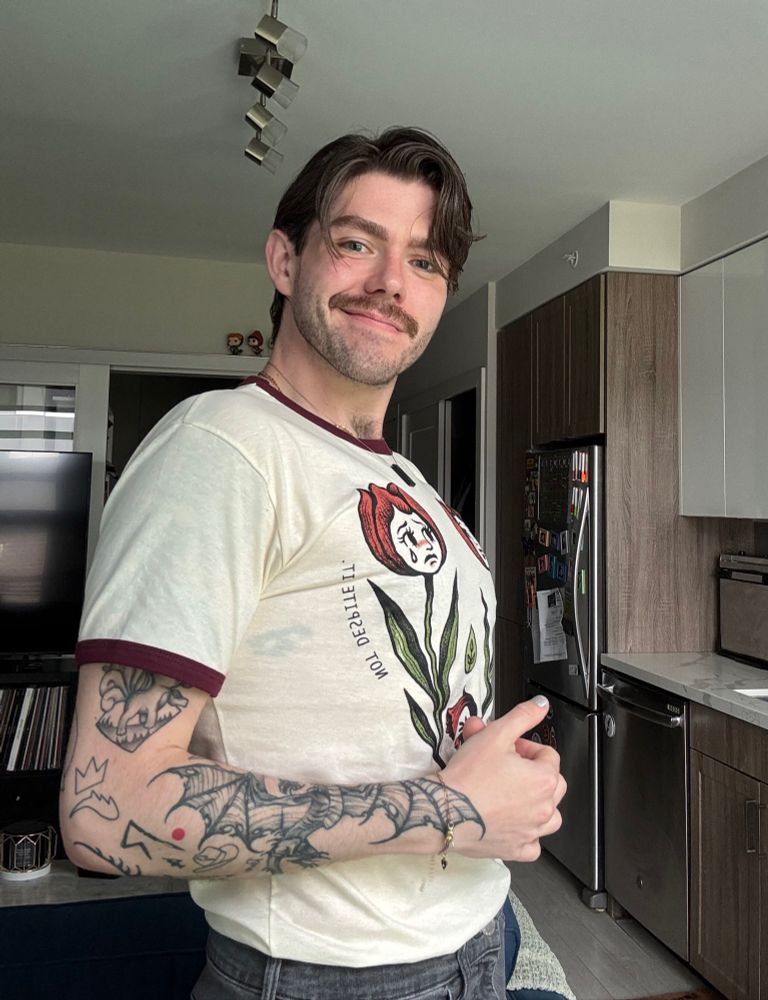 Me, from a side profile, wearing a graphic tee with red rings on the sleeves. My hair is swooping and in my face, brows nicely defined, and a good smile and dimple visible.