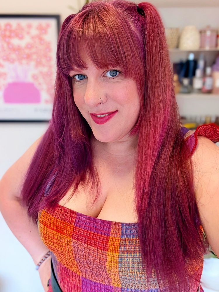 A woman with long pink hair and a bright checked dress smiles at the camera.