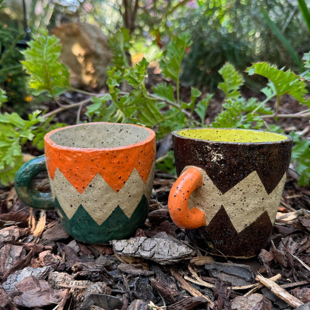 Two colourful pottery mugs displayed in a garden. Glaze design is a zigzag shape