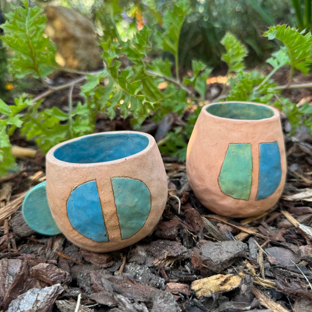 Two clay mugs photographed in a garden. Green and blue glaze, pale pink body 