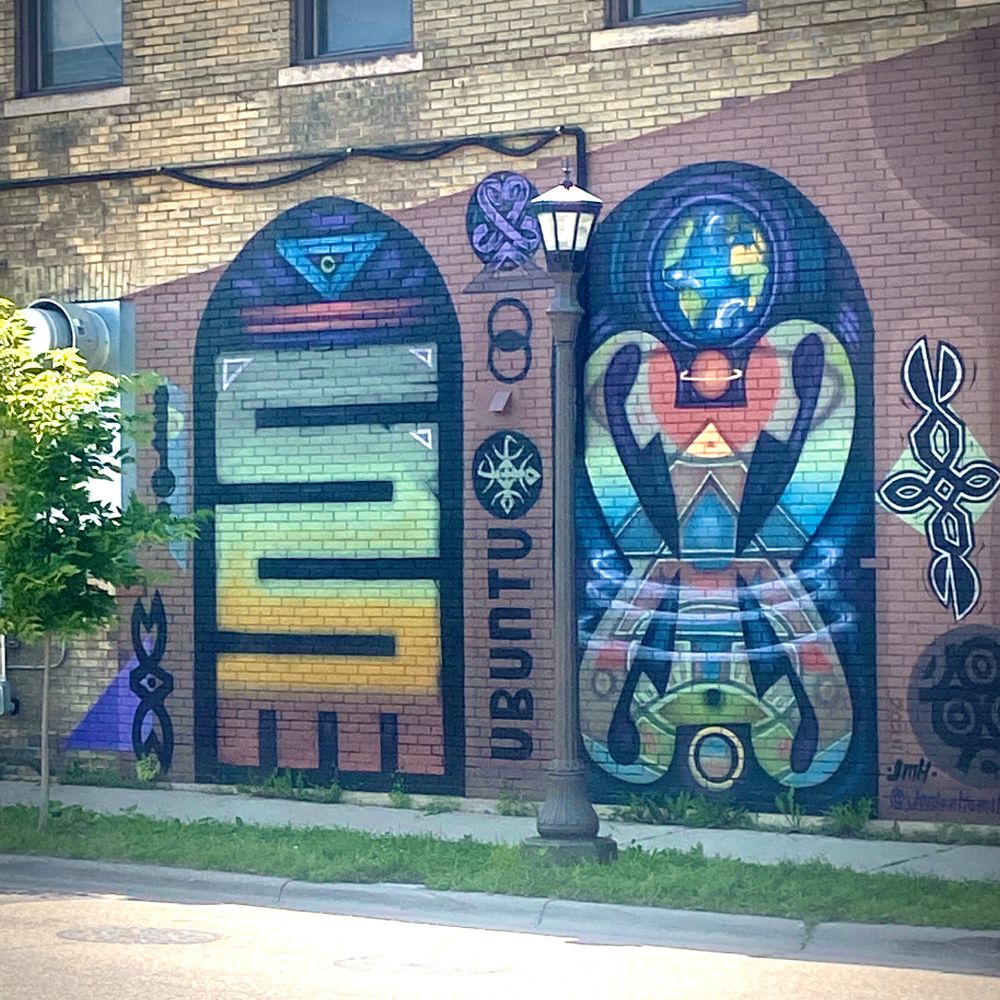 a large colorful mural painted on a brick building wall, next to a small tree and sunny street. two tall, dark, arch door shaped paintings with a earthy, sunset color scheme. each containing unique designs surrounded by geometric shapes. between them, printed vertically, the word ubuntu.
