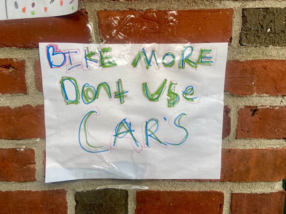 a handmade sign in a children's handwriting hanging on a brick wall. it reads "bike more dont use cars"